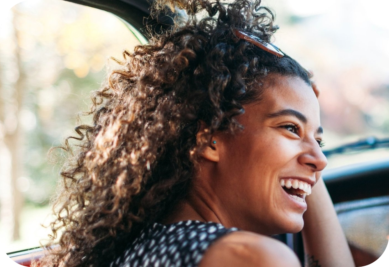 A woman with curly hair smiling while sitting in the passenger seat of a car.