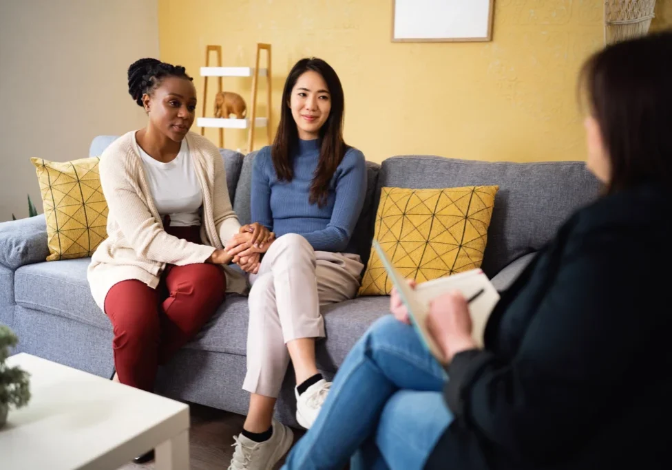 Two women sitting on a couch talking to another person.